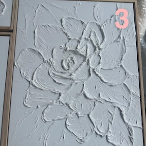 Op de afbeelding: Wit getextureerd bloemenkunstwerk op een vierkant canvas met een lichtbruine houten lijst. Het kunstwerk heeft een verhoogd, driedimensionaal ontwerp van een bloem. Het nummer "3" is zichtbaar in de rechterbovenhoek. Modern woondecoratie.