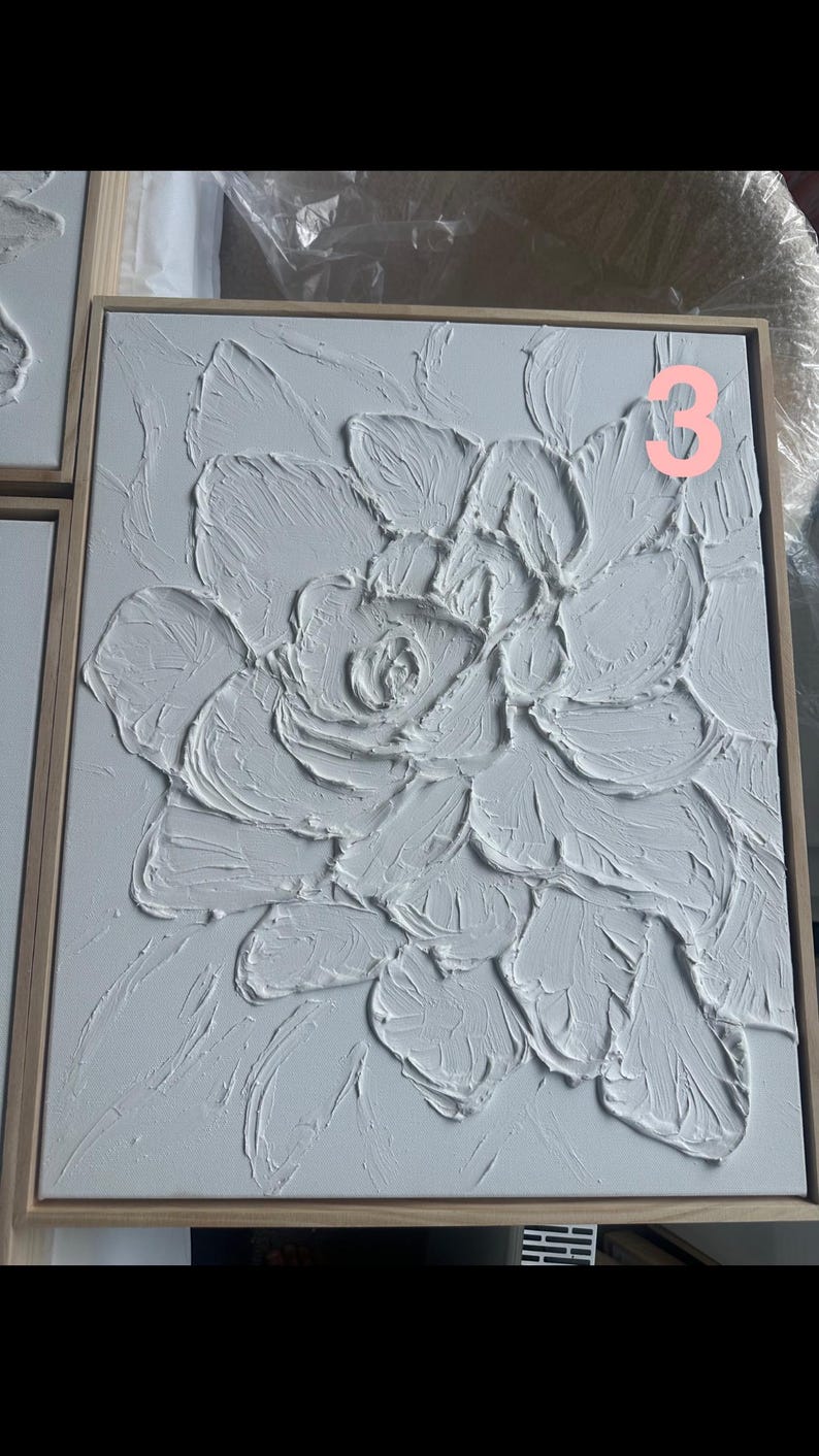 Op de afbeelding: Wit getextureerd bloemenkunstwerk op een vierkant canvas met een lichtbruine houten lijst. Het kunstwerk heeft een verhoogd, driedimensionaal ontwerp van een bloem. Het nummer "3" is zichtbaar in de rechterbovenhoek. Modern woondecoratie.