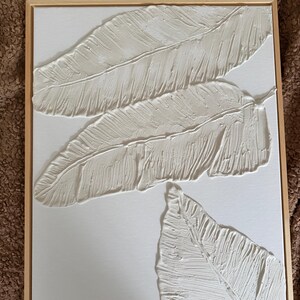 May include: Textured white feather art piece in a natural wood frame. The artwork features three dimensional feathers with detailed textures, creating a modern and minimalist aesthetic. The feathers are arranged on a white canvas.