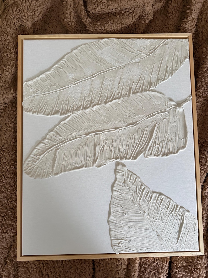 May include: Textured white feather art piece in a natural wood frame. The artwork features three dimensional feathers with detailed textures, creating a modern and minimalist aesthetic. The feathers are arranged on a white canvas.