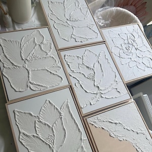 Op de afbeelding: Een verzameling witte, getextureerde bloemenkunstwerken in houten frames. Het kunstwerk toont verhoogde, driedimensionale ontwerpen van bloemen. De stukken worden gepresenteerd op een wit oppervlak, waarbij de ingewikkelde details en texturen van de kunst worden getoond.