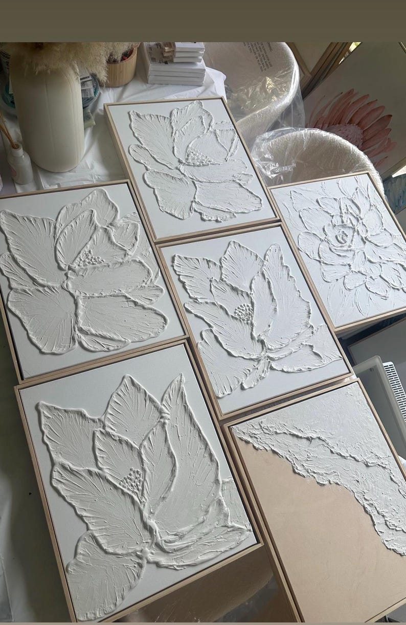 Op de afbeelding: Een verzameling witte, getextureerde bloemenkunstwerken in houten frames. Het kunstwerk toont verhoogde, driedimensionale ontwerpen van bloemen. De stukken worden gepresenteerd op een wit oppervlak, waarbij de ingewikkelde details en texturen van de kunst worden getoond.