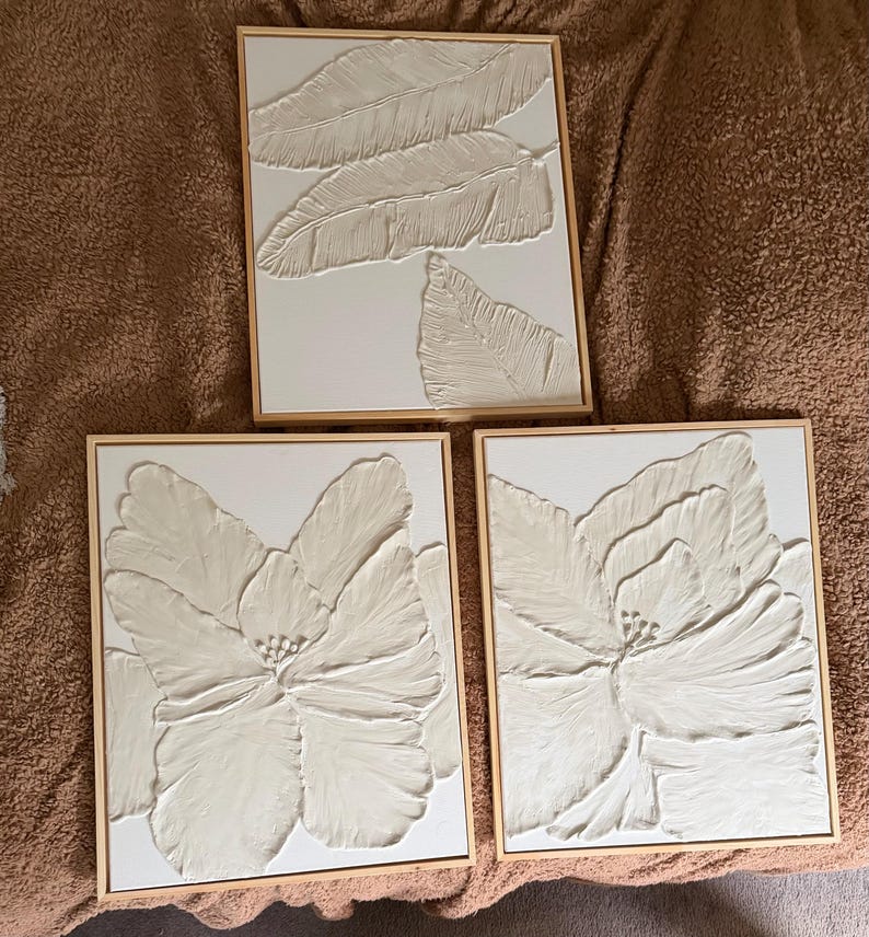 May include: Three framed wall art pieces. Two feature large, textured white flower designs, and one displays a textured white leaf design. All are set in light brown wooden frames, creating a cohesive set for home decor.