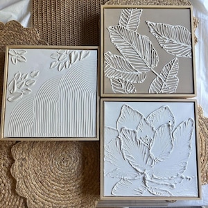 May include: Three framed canvas wall art pieces with white raised textured designs. The first piece features a white abstract design with a series of curved lines. The second piece features a white leaf design on a light brown background. The third piece features a white flower design on a white background.