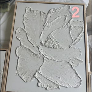 Op de afbeelding: Wit, getextureerd bloemenkunstwerk in een houten lijst. Het kunstwerk toont een grote, gestileerde bloem met gedetailleerde bloembladtexturen. Het nummer "2" is zichtbaar in de rechterbovenhoek. Het kunstwerk wordt op een wit oppervlak gepresenteerd.
