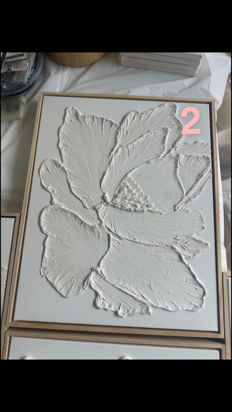 Op de afbeelding: Wit, getextureerd bloemenkunstwerk in een houten lijst. Het kunstwerk toont een grote, gestileerde bloem met gedetailleerde bloembladtexturen. Het nummer "2" is zichtbaar in de rechterbovenhoek. Het kunstwerk wordt op een wit oppervlak gepresenteerd.