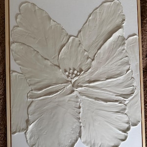 May include: A framed, textured, white floral art piece. The artwork features a large, stylized flower with detailed petals and a central stamen. The canvas is set in a natural wood frame, creating a modern, minimalist aesthetic.
