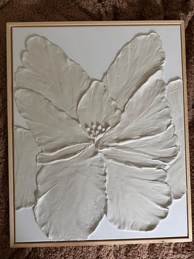 May include: A framed, textured, white floral art piece. The artwork features a large, stylized flower with detailed petals and a central stamen. The canvas is set in a natural wood frame, creating a modern, minimalist aesthetic.