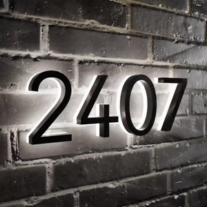 May include: Black metal house numbers with white LED lights, mounted on a brick wall. The numbers read "2407".