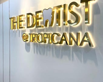 Può includere: Segno dorato con il testo "THE DENTIST @ TROPICANA" in lettere in rilievo. La parola "DENTIST" ha un'icona di dente stilizzata al posto della "T".