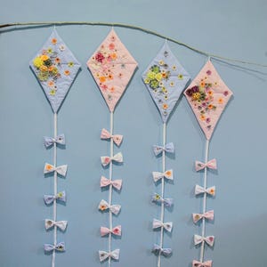 May include: Four blue and pink diamond-shaped kites with floral embroidery hang from a branch with white ribbon bows attached to each kite.