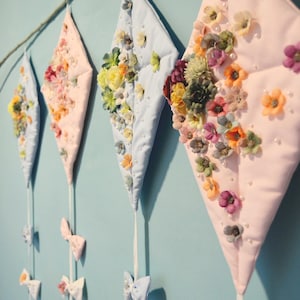 May include: Four diamond-shaped fabric kites with floral embellishments and white bows hanging from a branch on a light blue wall.