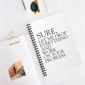 May include: White spiral-bound notebook with the text "SURE, LET ME DROP EVERYTHING AND WORK ON YOUR PROBLEM." Gold binder clip and sunglasses are also visible.