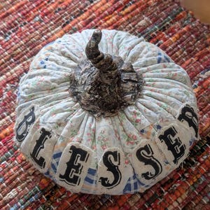 May include: A fabric pumpkin decoration featuring a light blue and white floral and plaid patchwork pattern. The pumpkin has a dark brown, textured stem and individual cream-colored fabric patches stitched on, each displaying a black letter spelling BLESSED.