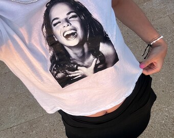 May include: A white cropped t-shirt with a black and white image of a woman laughing. The woman is wearing a necklace and has long dark hair. The shirt is being worn by a person with a black skirt and pink sneakers.