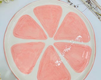 May include: A circular ceramic plate with a grapefruit slice design. The plate has a white base with six pink segments, a white border, and a pink rim. Decorative flowers are in the background, adding a natural touch.