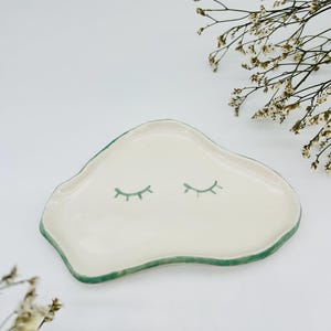 Puede incluir: Un plato de cerámica blanco con un borde verde y dos pestañas verdes pintadas en la superficie. El plato tiene forma de nube.