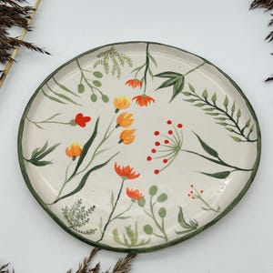 Puede incluir: Un plato de cerámica blanco con un borde verde presenta un diseño floral pintado a mano. El diseño incluye flores naranjas y rojas, hojas verdes y tallos.