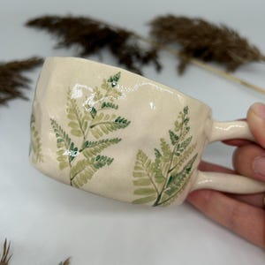 May include: A ceramic mug with a cream-colored glaze and a fern pattern in shades of green. The mug has a rounded shape and a handle.