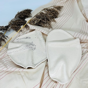 Op de afbeelding: Twee witte keramische serveerschalen met gespikkeld glazuur. De schalen liggen op een gestreepte stoffen achtergrond.