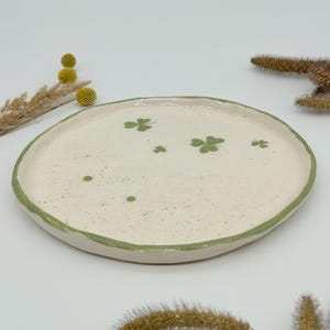 May include: A small, white ceramic dish with a green rim and green shamrock designs. The dish has a speckled finish.