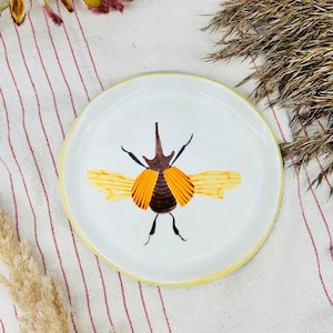 以下が含まれることがあります： 黒い持ち手と「But First, Coffee」のテキストが黒い太字で書かれた白いセラミックマグ。マグカップはぼやけた背景に置かれており、居心地の良い環境を示唆しています。