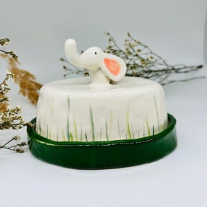 Può includere: Una statuetta di elefante in ceramica bianca con orecchie rosa, seduta su una base in ceramica verde e bianca con un design a erba.