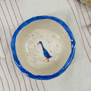 May include: A handmade ceramic bowl with a blue rim and a whimsical duck illustration. The interior is off-white with a blue duck and red raindrop accents. The bowl is approximately 6 inches in diameter.