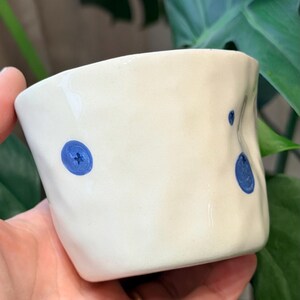Pode incluir: Uma caneca de cerâmica branca feita à mão, com uma forma ligeiramente irregular. A caneca é decorada com três desenhos de mirtilos azuis pintados à mão. A caneca é segurada em uma mão, com um fundo de folhas verdes.