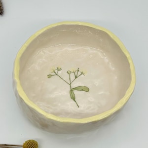Puede incluir: Un cuenco de cerámica hecho a mano con interior crema y borde amarillo pálido. El cuenco presenta un delicado diseño floral en el centro. Perfecto para servir alimentos o como pieza decorativa.