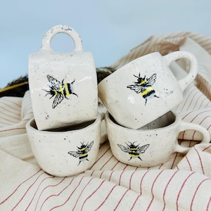 May include: Set of four white ceramic mugs with black speckled glaze. Each mug features a hand-painted black and yellow bumblebee design.