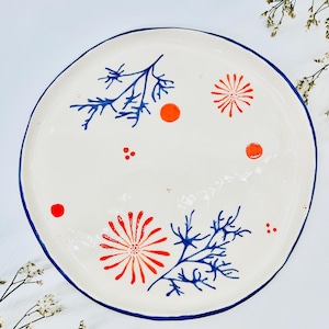 Può includere: Un piatto in ceramica bianca con un bordo blu e disegni floreali arancioni e blu dipinti a mano. Il piatto presenta fiori e rami stilizzati.