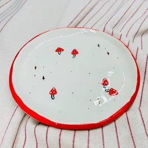 May include: A white ceramic plate with a red rim, decorated with small red and white mushroom illustrations and black star-shaped accents. The plate is round and sits on a white and red striped fabric.