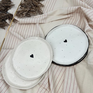Peut inclure: Deux assiettes en céramique blanche avec des pois noirs et un motif de cœur noir. Les assiettes sont empilées l'une sur l'autre et reposent sur un tissu rayé.