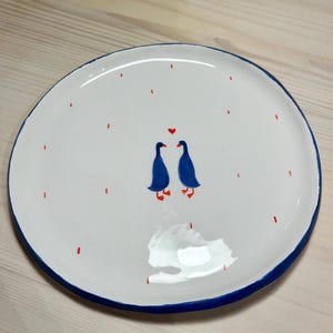 Pode incluir: Prato de cerâmica branco com borda azul. O prato apresenta um design caprichoso de dois pássaros azuis voltados um para o outro, com um coração vermelho acima deles. Traços verticais vermelhos são espalhados ao redor dos pássaros. Uma peça decorativa para a casa.