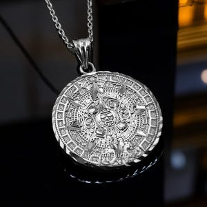 May include: Silver-toned pendant necklace featuring a detailed Aztec calendar design. The circular pendant is suspended from a delicate chain. The intricate pattern includes various symbols and shapes, creating a detailed and eye-catching piece of jewelry.