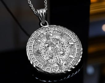May include: Silver-toned pendant necklace featuring a detailed Aztec calendar design. The circular pendant is suspended from a delicate chain. The intricate pattern includes various symbols and shapes, creating a detailed and eye-catching piece of jewelry.