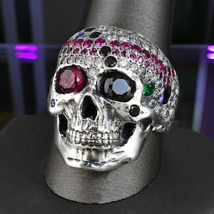 May include: A silver skull ring with a detailed design. The skull's head is adorned with a variety of colorful gemstones, including red, black, and clear stones. The ring is displayed on a black stand.