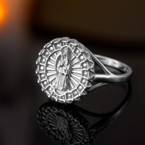 May include: A silver ring featuring a detailed depiction of a robed figure within a circular frame. The ring's design includes a sunburst pattern around the central image. The ring is set against a dark, reflective surface.