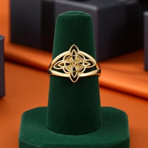 May include: A gold-colored ring with an intricate Celtic knot design, displayed on a green velvet ring holder. The ring features a central knot with four surrounding loops. The background includes black gift boxes with black ribbons.