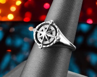 May include: A silver compass ring with a black background. The ring is on a black ring display stand. The background is out of focus and features red and blue lights.