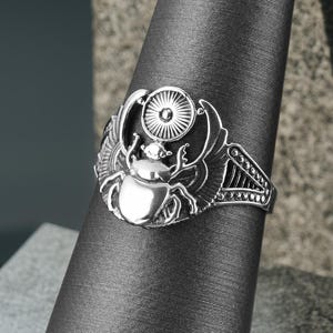 May include: A silver ring featuring an Egyptian scarab beetle design. The ring has intricate details, including wings and a sun disc. The ring is displayed on a gray fabric cone, showcasing its design and craftsmanship.