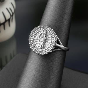 May include: A silver ring with a detailed religious design, displayed on a black ring holder. The ring features a central figure surrounded by a circular pattern. A decorative skull is visible in the background.