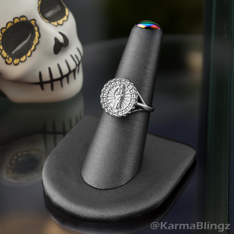 May include: A silver ring with a detailed religious design, displayed on a black ring holder. The ring features a central figure surrounded by a circular pattern. A decorative skull is visible in the background.