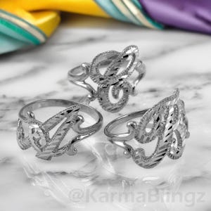 May include: Three silver-colored initial rings, each with a different letter design. The rings are displayed on a reflective surface with a marble pattern. The letters have a textured, patterned appearance.