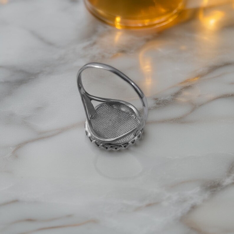 May include: A silver ring with a large, oval-shaped setting. The setting is surrounded by small, decorative stones. The ring's band curves up to form a loop above the setting. The ring is displayed on a white and gray marble surface.