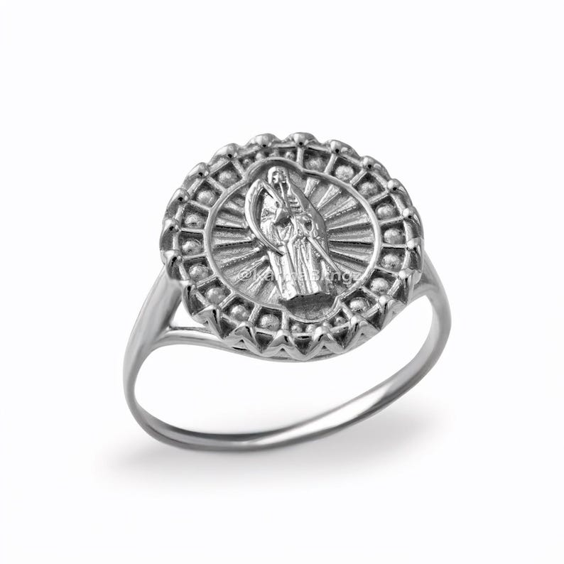 May include: A silver ring featuring a detailed central design of a robed figure with a scythe, surrounded by a sunburst pattern within a decorative, circular frame. The ring has a polished finish and a simple band.