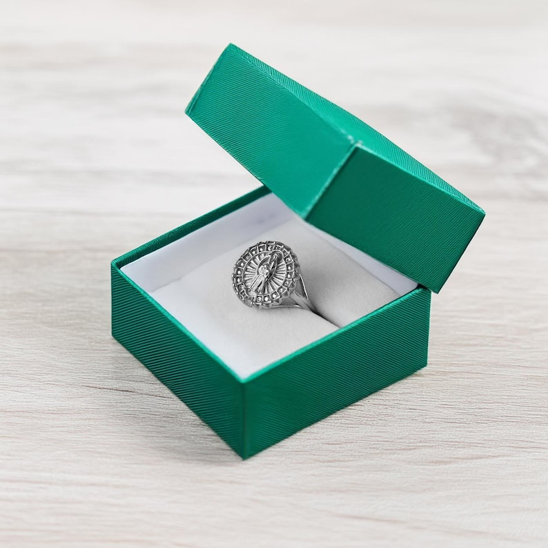 May include: A silver ring with a circular, ornate design featuring a central element and surrounding details, presented in an open, emerald green jewelry box with a white interior.