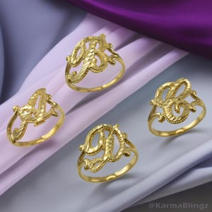 May include: Four gold-colored initial rings, each with a different letter: A, B, and D. The rings have a textured, ornate design. The rings are displayed on a light purple and gray background.