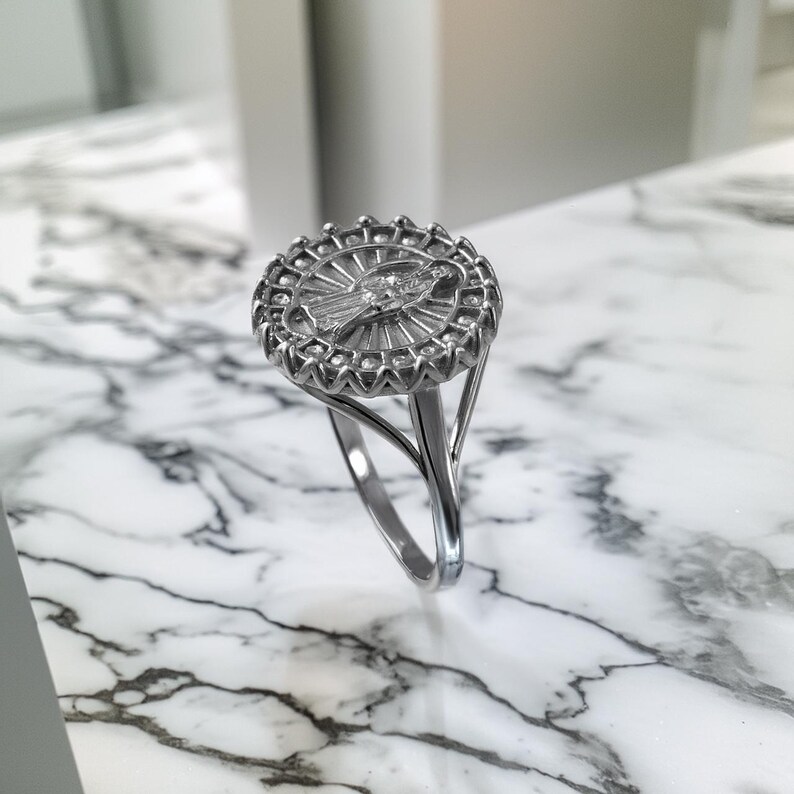 May include: Silver-toned ring featuring a detailed religious icon within a circular frame. The ring's design includes a central figure surrounded by radiating lines and a decorative outer edge. The band splits into two graceful arcs, creating an open design.